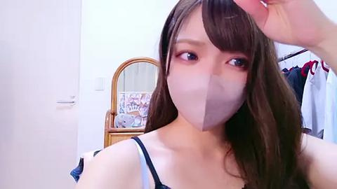 Media: Video of a young Asian woman with long brown hair and pale skin, wearing a black and white bra, adjusting her hair in a small, tidy bedroom with a wooden vanity and a mirror.