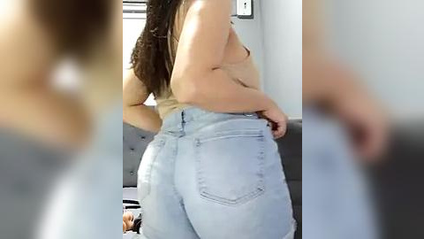 Media: A video of a woman in light blue jeans, showing her bare back and side, standing in a modern, grey-toned room.