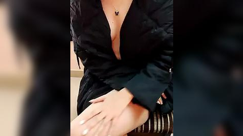 Media: A video of a person sitting, wearing a black, open-front dress revealing cleavage, with a black butterfly necklace, hands resting on knees.