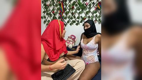 Media: A video of two women in a room, one in a red headscarf and beige pants, the other in a black headscarf and pink lingerie, sitting on a bed with a colorful floral backdrop.