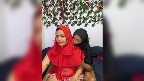 Media: Video: A young woman in a red hijab and floral-patterned skirt sits on a bed, with another woman in a black hijab holding her from behind. The background features a decorative wall with green leaves and colorful flags.