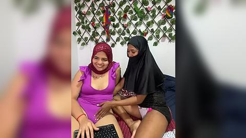 Media: A video shows two women: one in a purple dress, the other in a black burqa, sitting on a bed. The background features decorative plants and a white wall.