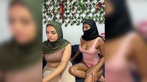 Media: Video of two women in a room: one in a green hijab and beige top, the other in a pink hijab and top, both sitting on a dark sofa.