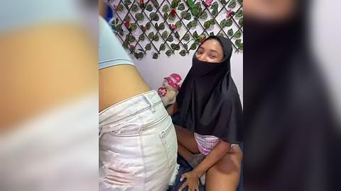 Media: Video of a woman in a black hijab, kneeling with a pink mask on, licking a man's exposed penis through his pants. Background features a floral-patterned wall and a white ceiling.