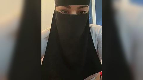 Media: Video of a person with a black face veil, brown eyes, and a white shirt, standing behind bars, with blurred bars in the foreground.
