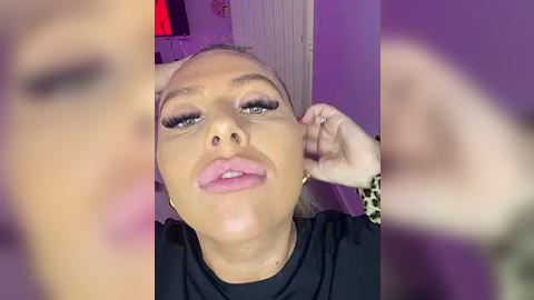 Media: A close-up video of a light-skinned woman with blonde hair, wearing dramatic makeup, including long eyelashes and pink lipstick, adjusting her hair. The background is blurred, showing purple walls and a partially visible red object.