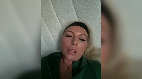 Media: Video of a blonde woman in a green jacket, lying on a textured white ceiling, eyes closed, mouth slightly open.