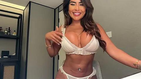 Media: Video of a smiling, dark-haired woman in white lace lingerie, standing in a modern bedroom with light gray walls and a black-framed mirror.