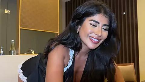 Media: Video of a smiling South Asian woman with long black hair, wearing a black lace dress, in a modern room with beige and brown walls.