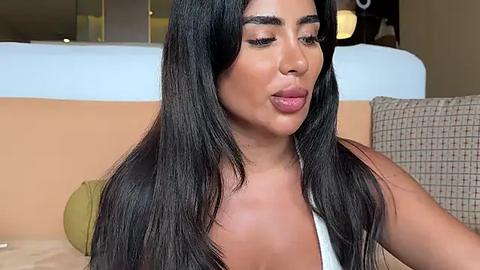 Media: Video of a young woman with long, straight black hair, wearing a white top, sitting on a beige couch. She has a medium tan skin tone, full lips, and subtle makeup. Background shows a modern, neutral-toned living room.