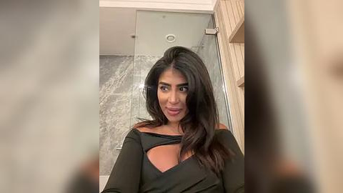 Media: A video of a woman with long, dark hair, wearing a black off-shoulder top, standing in a modern bathroom with glass shower doors and wooden accents.