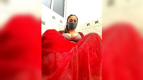 Media: Video of a woman with a medium skin tone, wearing a red mask, red blouse, and gray face mask, seated on a red pleated blanket, indoors with beige tiled walls.