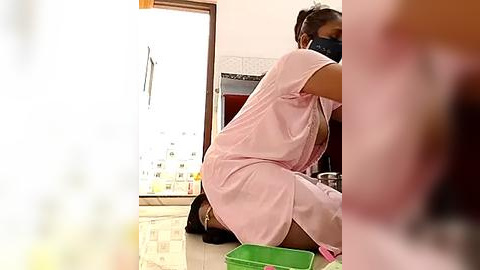 Media: Video of a nurse in pink scrubs, wearing a face mask, squatting beside a green trash can, in a brightly lit, clean room with white walls and tiled floor.