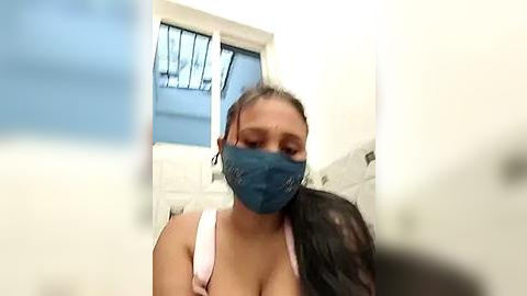 Media: Video of a woman with light brown skin, dark hair tied back, wearing a blue surgical mask and white tank top, sitting in a brightly lit bathroom with white walls and a window.