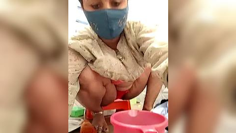 Media: A video of a woman with a blue mask and a beige top, exposing her breasts, bending over a red and pink toy box.