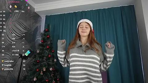 Media: Video of a young woman in a gray striped sweater, red Santa hat, and red gloves, dancing joyfully in front of a decorated Christmas tree, with teal curtains and a black inflatable Santa on the left.