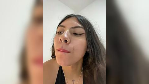 Media: Video of a young Latina woman with long, dark hair and light skin, wearing a black top, and a nose ring. She has a neutral expression. Background is a plain, white-walled room.