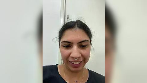 Media: A video of a smiling, dark-haired woman with medium skin tone, wearing a black top, in a white-walled room. Her nose is pierced with a septum ring.