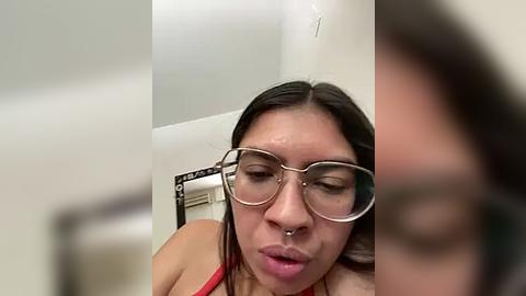 Media: Video of a young woman with long dark hair, wearing oversized glasses, a nose ring, and a red top, sitting on a white toilet. The background is blurred.