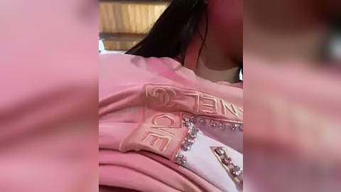 Media: Video of a woman with long black hair wearing a pink Chanel top with \"CHANEL\" embroidered in gold. She is lying on a white surface, with a blurred background of wooden panels.