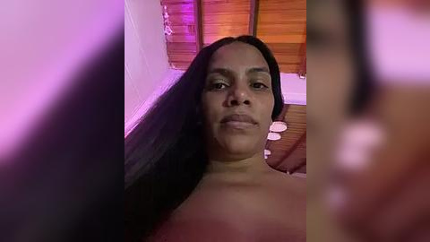 Media: A video of a topless woman with long, straight black hair, light brown skin, and a serene expression. She is in a dimly lit room with wooden walls and purple lighting, her body partially obscured by out-of-focus figures.