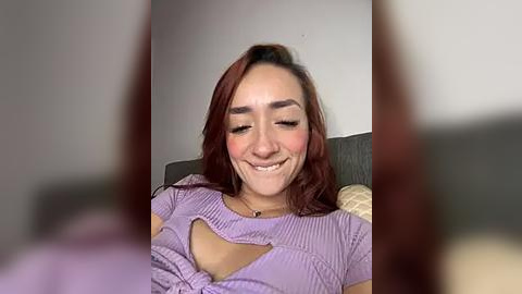 Media: Video of a young woman with light skin, reddish-brown hair, and a medium build, wearing a purple, ruffled crop top, smiling, seated on a grey couch in a dimly lit room.