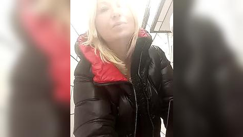 Media: A video of a blonde woman in a black puffy jacket with a red lining, standing in a tiled subway station, slightly blurred.