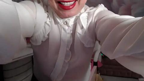 Media: Video of a smiling person wearing a white, frilly blouse, possibly in a kitchen setting. The background is blurred, with hints of appliances and countertops. The image is casual and candid.