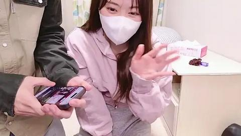 Media: Video of a young Asian woman with long brown hair, wearing a white mask and light pink jacket, holding a phone and a small purple box of pills. Background includes a white wall and a green chair.