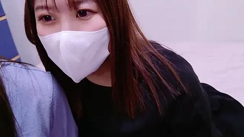 Media: Video of an East Asian woman with straight, long brown hair, wearing a white face mask and a black top, lying on a light blue pillow in a clean, minimalistic room.