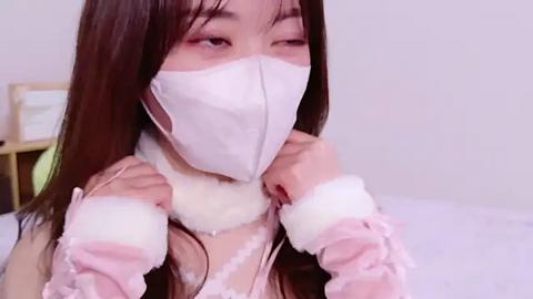 Media: A video of an East Asian woman with long brown hair, wearing a white face mask, a pink lace top, and fluffy white sleeves, adjusting a pink ribbon on her chest. The background is a white wall and a wooden headboard.