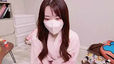 Media: Video of an East Asian woman with long, straight brown hair, wearing a white face mask and pink pajamas, kneeling on a grey carpet in a cluttered room with a toy box, stuffed animals, and books.