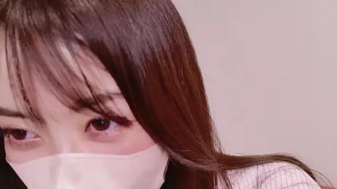 Media: Video of an Asian woman with straight, shoulder-length brown hair, wearing a white surgical mask, partially covering her face, with large, expressive brown eyes visible. The background is a plain, light beige wall.