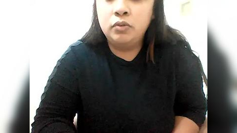 Media: Video of a middle-aged woman with medium skin tone and straight black hair, wearing a black long-sleeve top, looking serious with pursed lips, against a blurred white background.