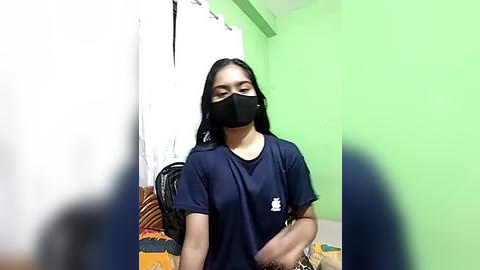 Media: Video of a young woman with long black hair, wearing a black mask, navy t-shirt, and sitting on a bed with green walls and white curtains in the background.