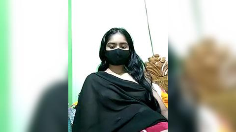 Media: Video of a South Asian woman with long black hair, wearing a black mask, black sari, and red blouse, seated in a green and white room with a wooden chair.