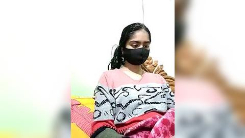 Media: Video of a young woman with dark skin, wearing a black face mask, pink and white striped sweater, and a red blanket, sitting against a plain white background.