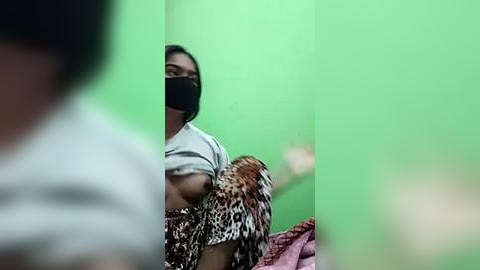 Media: Video of a woman in a leopard-print dress, wearing a face mask, sitting in a dimly lit room with a green wall, holding a small bird.