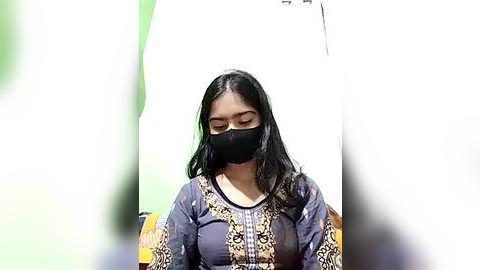 Media: A video of a woman with long black hair, wearing a black face mask and a colorful, patterned long-sleeve top, standing in a brightly lit room with green walls.