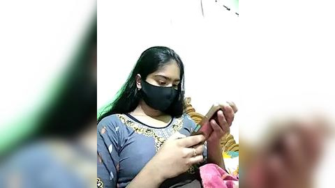 Media: Video of a South Asian woman with medium skin tone, wearing a blue dress with gold embroidery, black mask, and gloves, applying a vaccine to a child.