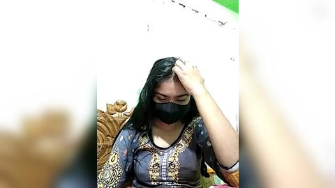 Media: Video of a South Asian woman with long black hair, wearing a face mask and a colorful, patterned blouse, sitting on a wooden chair, touching her head in distress. Background shows a white wall with a green border and a string of lights.