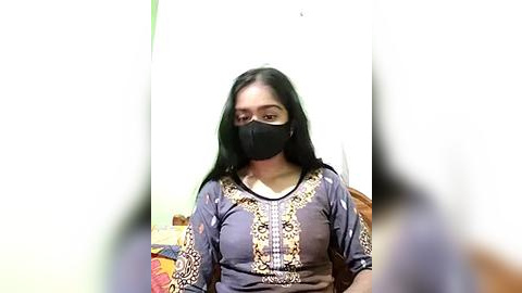 Media: Video of a South Asian woman with long black hair, wearing a black face mask, purple embroidered shirt, and sitting indoors against a plain white wall.