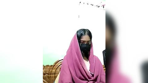 Media: A video of an Indian woman with a brown complexion, wearing a pink sari and black mask, standing in front of a beige wall.