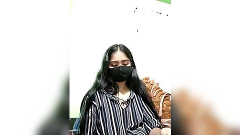 Media: Video of a young woman with long black hair, wearing a black face mask, a striped shirt, and sitting indoors on a wicker chair.