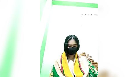 Media: Video of a woman in traditional South Asian attire, wearing a yellow and green shawl over a white shirt, black mask covering her nose and mouth, and a green wall behind her.