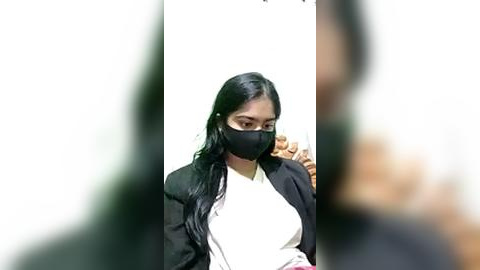 Media: Video of a woman with long black hair, wearing a black mask, white shirt, and black blazer, holding a phone, blurred background, indoor setting.