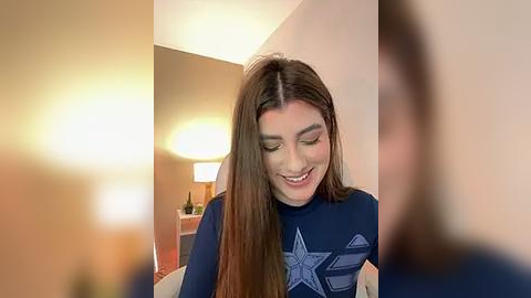 Media: Video of a young woman with long brown hair, wearing a navy blue sweater, smiling, with a blurred background featuring a modern, warmly lit room with a lamp.