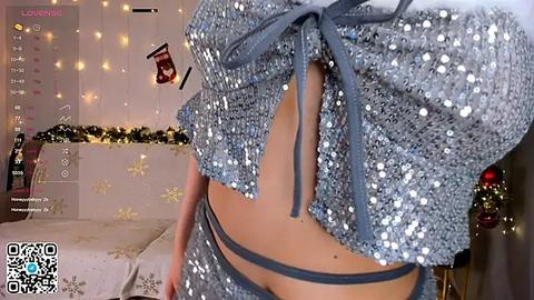 Media: Video of a woman in a sparkly gray crop top and matching skirt, with a festive background featuring holiday lights, a wrapped gift, and a Christmas tree.