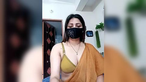 Media: Video of a South Asian woman with medium skin tone, wearing a mustard-yellow sari, black face mask, and a light blue bra, in a simple indoor setting with a green plant in the background.