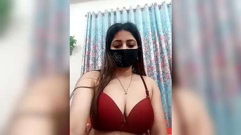 Media: Video of a South Asian woman with medium complexion, long black hair, wearing a red bra, black face mask, and floral-patterned curtains in the background.